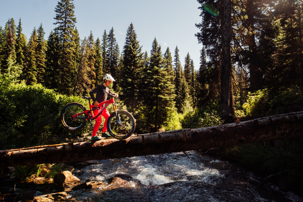 A biker crosses a river with her bike by walking across a log. 
